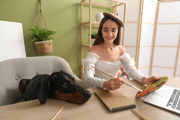 Cute Doberman dog and beautiful young happy woman in beret with laptop holding paints palette at...