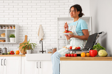 Beautiful young happy African-American woman with plate of vegetables near modern electric grill sitting in kitchen