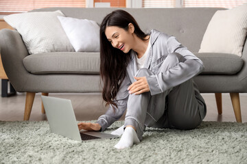 Fototapeta premium Young woman in grey pajamas working with laptop on carpet at home