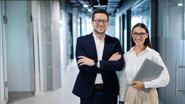 Portrait of two business colleagues posing with crossed arms and smiling at camera in company hall, standing together at contemporary office. Coworking concept