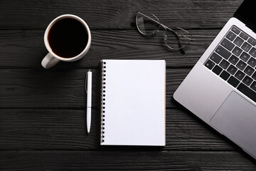 Open blank notebook, pen, coffee, laptop and glasses on black wooden table, flat lay