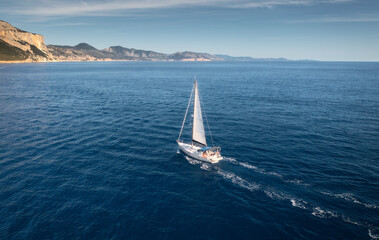 Plakat Aerial view of floating sailboat and rocky sea coast at summer sunny day. Top drone view of yacht, mountains, trees and blue water. Yachting in Sardinia, Italy. Tropical seascape with boat in ocean
