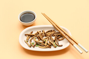 Plate with fried maggots and bowl of soy sauce on orange background