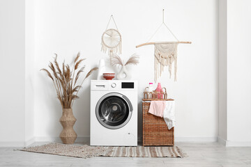 Modern washing machine with laundry basket and pampas grass near white wall in bathroom