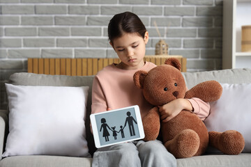 Sad girl with toy bear and family figure sitting on sofa at home. Adoption concept