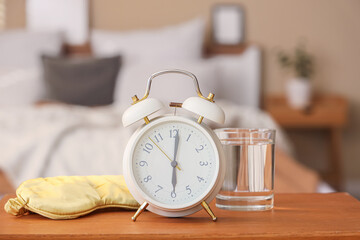 Fototapeta na wymiar Alarm clock with glass of water and sleep mask on bedside table in bedroom, closeup