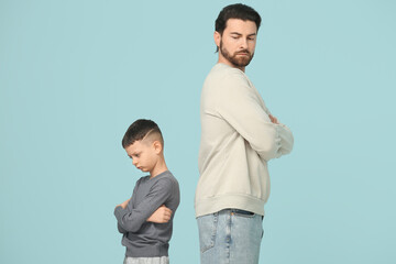 Young father and his little son with crossed arms not looking at each other after squabbling on blue background