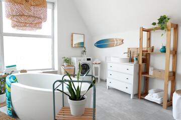 Interior of light bathroom with bathtub, washing machine, big window and surfboard
