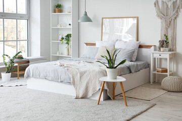 Interior of light bedroom with cozy bed, bedside table, houseplants and shelving unit