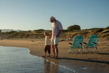 Grandfather holding grandson's hand while walking barefoot along the beach shore, enjoying quiet family moment during golden hour on a summer evening.