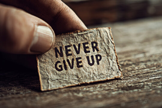 A close-up image of a hand holding a message card that reads "NEVER GIVE UP," symbolizing persistence, motivation, and resilience in the face of challenges.