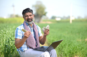 Indian farmer showing money or currency notes while working on laptop at crop field. concept of...