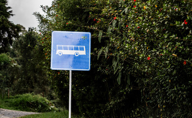Señal azul de parada de bus ubicada en zona rural rodeada de vegetación y flores, bajo cielo nublado