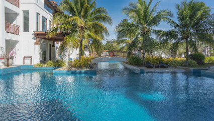 Blue outdoor swimming pool. Pedestrian bridge over the water. Lifebuoy on the railing. Deck chairs by the water. Tropical vegetation, palm trees. A fragment of a building nearby. Azure sky.Philippines