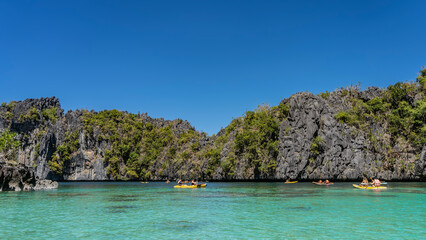 Obraz premium Tourist canoes sail through the beautiful turquoise lagoon surrounded by sheer cliffs. Green tropical vegetation on steep slopes. Clear blue sky. Copy space. Philippines. Palawan. Big Lagoon.