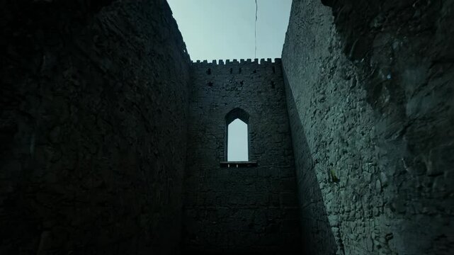 Zoom out from a small, barred window of an old castle dungeon to reveal the vast, oppressive stone walls, lit by faint ambient light