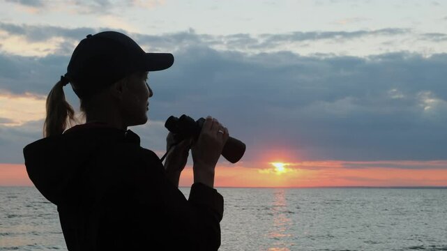 Silhouette of a woman in a cap looks through binoculars against the background of sky and sea at sunset. Portrait of a female tourist using binoculars.