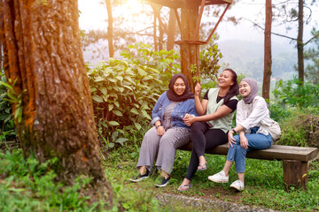 Fototapeta premium Three Asian Indonesian Women Laughing Together on Bench in Forest Park Setting