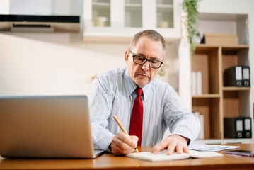 Naklejka premium senior businessman in red tie working on laptop at desk in modern kitchen home office. Remote work, corporate mood