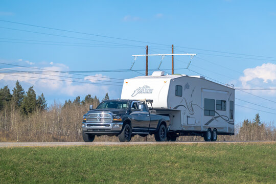 Calgary, Alberta, Canada. May 9, 2025. A black Dodge RAM 2500 truck tows a white 2001 Lakota 5th Wheel trailer along a road, with power lines overhead and a grassy field beside.