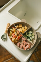 Divided silicone toddler plate on a highchair tray filled with diced cucumber, cheese cubes, colorful pasta spirals and sliced sausage