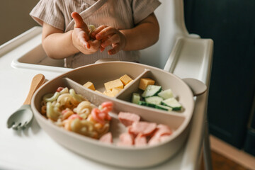 A toddler’s chubby hands grasp colorful pasta and cheese cubes from a divided plate during mealtime