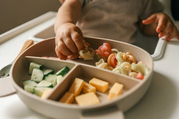 A toddler’s chubby hands grasp colorful pasta and cheese cubes from a divided plate during mealtime