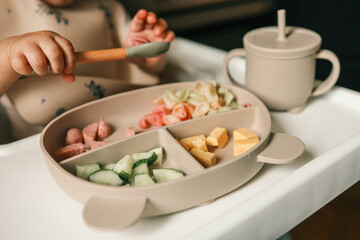 A toddler's chubby hands grasp colorful pasta and cheese cubes from a divided plate during mealtime