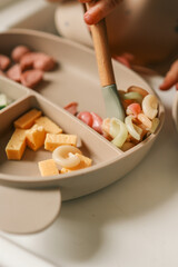 A toddler’s chubby hands grasp colorful pasta and cheese cubes from a divided plate during mealtime