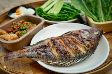 A traditional Thai meal is beautifully arranged on a round bamboo tray. At the center is a perfectly grilled whole fish, served alongside a variety of fresh vegetables, cucumbers, and boiled eggs.