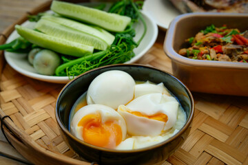 A traditional Thai meal is beautifully arranged on a round bamboo tray. At the center is a perfectly grilled whole fish, served alongside a variety of fresh vegetables, cucumbers, and boiled eggs.