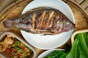 A traditional Thai meal is beautifully arranged on a round bamboo tray. At the center is a perfectly grilled whole fish, served alongside a variety of fresh vegetables, cucumbers, and boiled eggs.