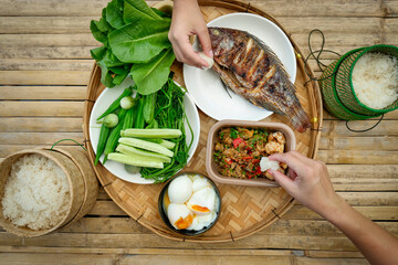 A traditional Thai meal is beautifully arranged on a round bamboo tray. At the center is a perfectly grilled whole fish, served alongside a variety of fresh vegetables, cucumbers, and boiled eggs.