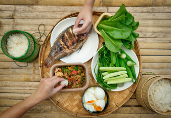 A traditional Thai meal is beautifully arranged on a round bamboo tray. At the center is a perfectly grilled whole fish, served alongside a variety of fresh vegetables, cucumbers, and boiled eggs.