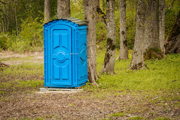 Vivid blue plastic portable toilet stands on a wooden base among tall trees in a forest clearing, surrounded by grass and fallen leaves in spring.