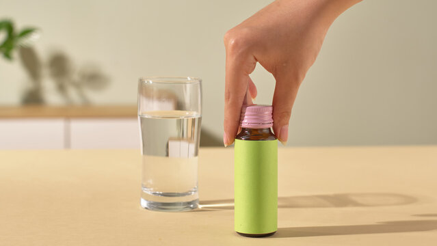 Frontal shot of a hand attempting to pick up a glass bottle of collagen. The bottle has a green body and purple lid, and a glass of water sits on the table. Natural light shines on the objects.