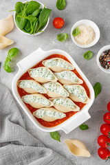 Raw conchiglioni pasta stuffed ricotta cheese and spinach with tomato sauce in a white baking dish on a gray concrete background.