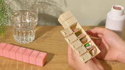Two hand holding a beige personal daily pill box, inside each compartment are colorful pills, arranged according to the doctor's prescription. Another pink pill box is placed on the table.