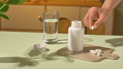 A hand is picking up a pill, some capsules placed on the tray, next to it is an open medicine bottle, displayed on a light brown tray. A glass of pure white water and a pot of green plants.