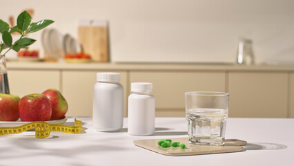 A glass of purified water sits on a tray with green pills. On the table are unlabeled pill bottles along with yellow. Body measuring tape and fresh apples. The concept of medicine in the kitchen.