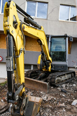 Compact excavator at a construction site near a modern building. Heavy machinery used for urban development and renovation works. Building facade in the background, rubble and dirt in the foreground.