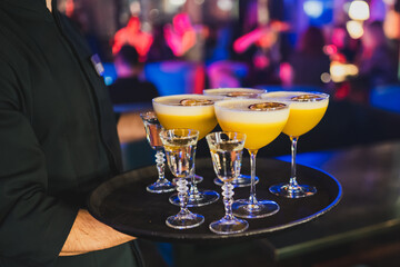 A waiter in a dark uniform holds a tray with four yellow cocktails garnished with dried fruit and four clear shot glasses. background is a vibrant, colorful bar scene with blurred lights and people.