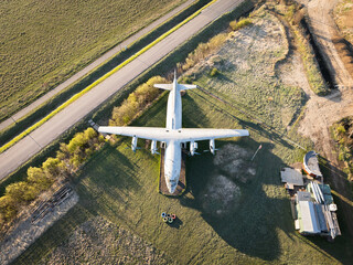 Aerial photography of an old Soviet cargo plane parked in a green field next to a road in a rural area.