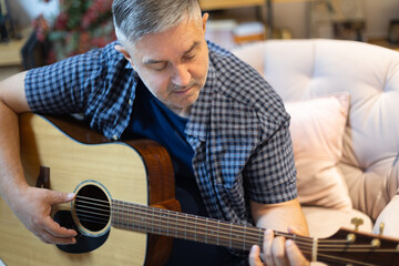 Portrait of a mature man sitting in a cozy chair at home, focused on playing an acoustic guitar. A...