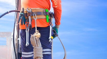 Midsection of electrician with safety belt and work tool is climbing to installing electrical equipment on electric power pole against blue sky background