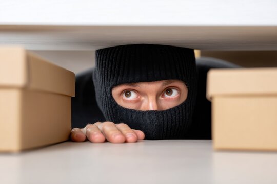 Thief in balaclava hiding under desk, looking at cardboard boxes, stealing.