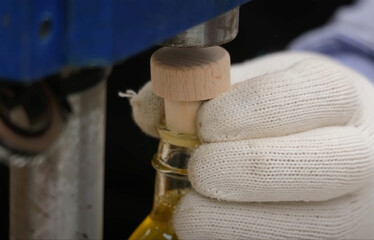 Wooden cap being securely pressed onto a glass bottle by a capping machine, with a gloved hand holding the bottle during a clean, safe production process.