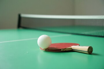A red paddle rests on a green table tennis surface, a white ball beside it, near a blurred net.