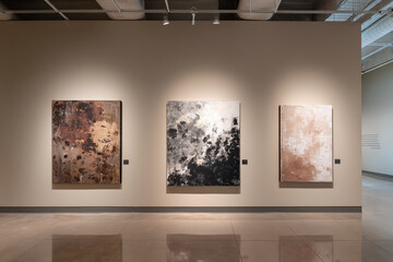 Three abstract paintings displayed on beige wall in art gallery showcase, featuring brown, gray and white color palettes, creating a modern and minimalist aesthetic