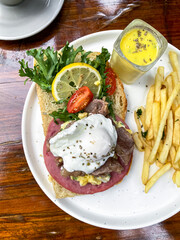 Side plate shot of open sandwich featuring half boiled egg, smoked beef, and fries in a bright presentation.
